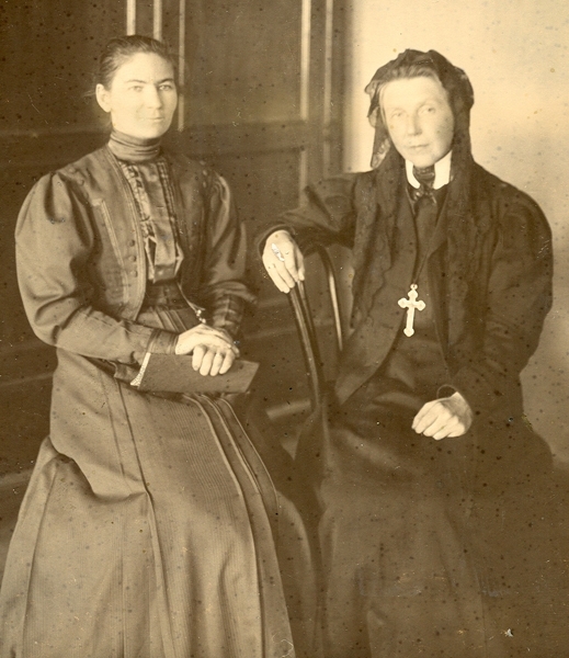 Na zdjęciu dwie kobiety w sukniach pozują do zdjęcia. 
