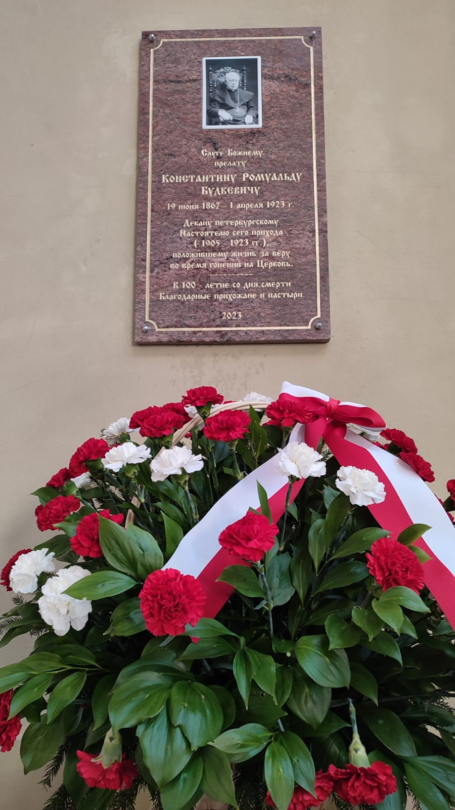 Ściana budynku. Na niej zawieszona marmurowa tablica pamiątkowa. Poniżej duży bukiet kwiatów z wstęgą w barwach polskiej flagi. 